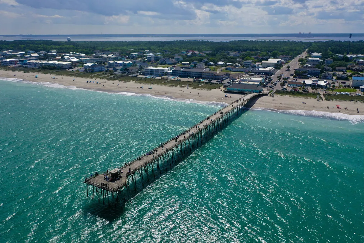 An expansive exterior scene of a pier extending into the ocean with the beach and town visible in the background. The area is bustling yet relaxed, presenting a quintessential beach community vibe ideal for sunset strolls and exploration.
