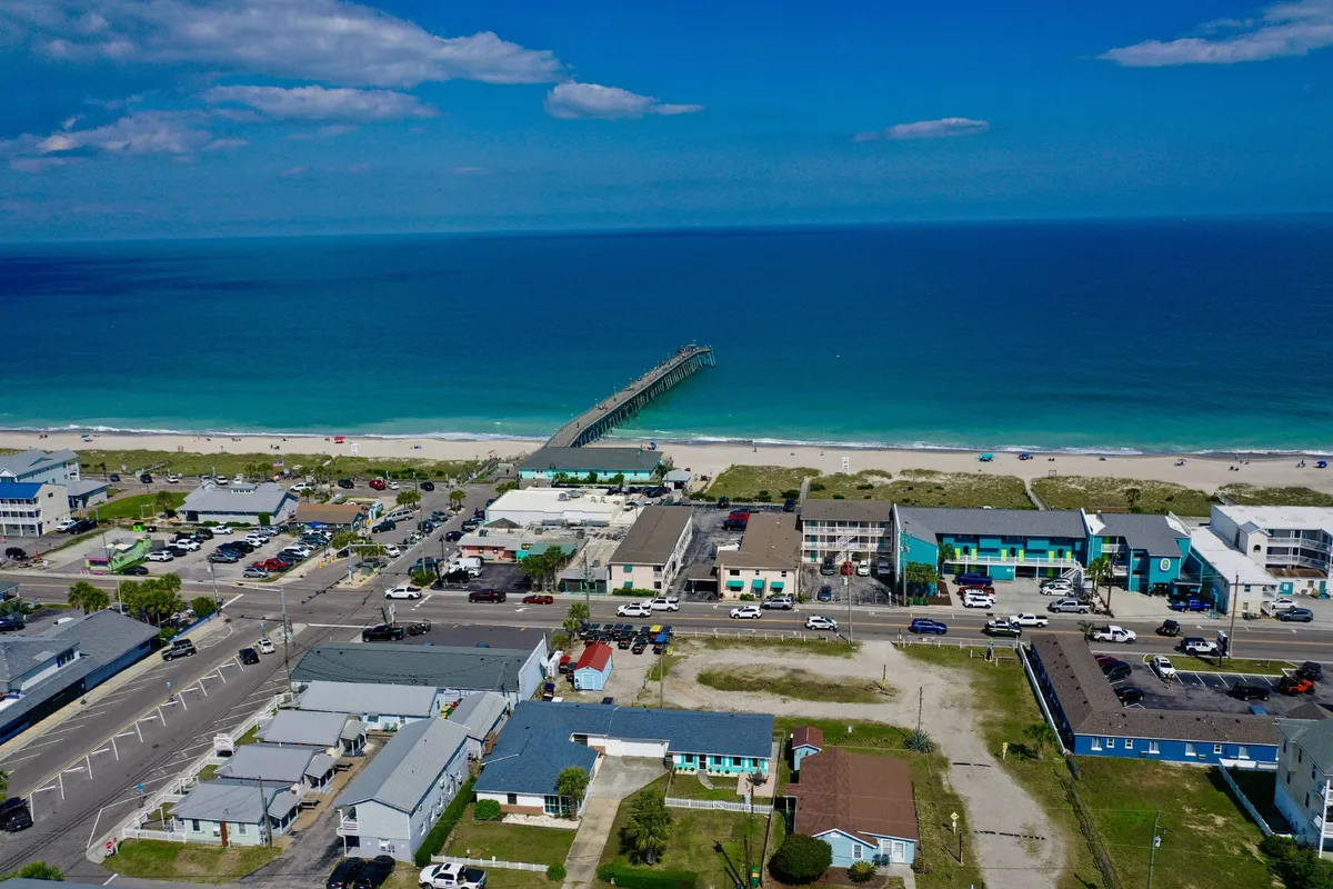 A panoramic view that captures the pier, ocean, sandy beach, and nearby buildings, with people engaging in beachcombing, fishing, and enjoying local dining and shopping. The inviting mood showcases the vibrancy and walkability of the area.