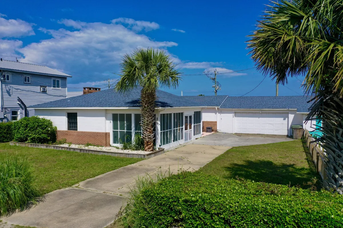 The image showcases the charming exterior of Lorraine's Beach Cottage, framed by swaying palm trees and a welcoming front entrance. The driveway and garage provide convenient parking, while the inviting porch hints at relaxing mornings and sunset views. The overall scene evokes a sense of relaxation and coastal living, perfect for families or groups seeking a beach getaway.