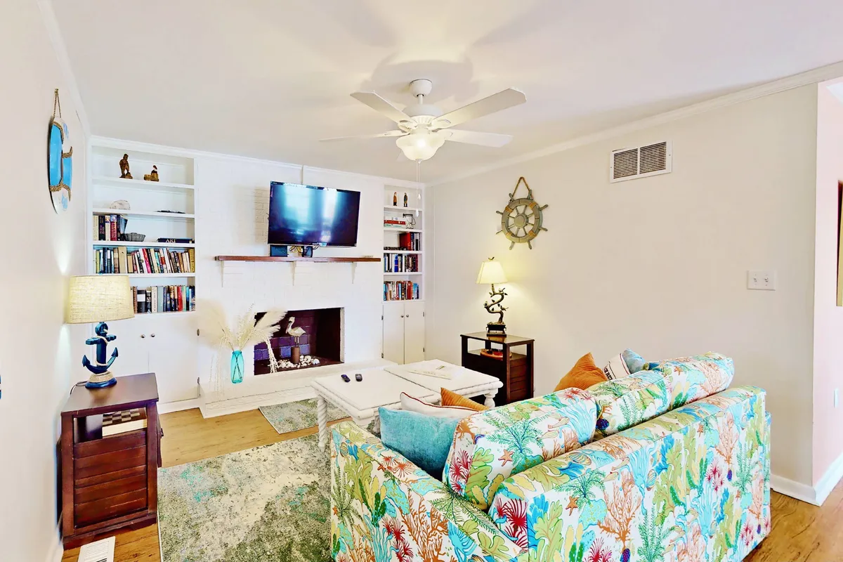 This image showcases a bright and cheerful living room with a colorful sofa, a cozy fireplace, a television, and a bookshelf. The room is bathed in natural light, highlighting the inviting seating area that's perfect for movie nights or socializing. The overall decor is breezy and coastal, with thoughtful touches that make the space feel both lively and comfortable.