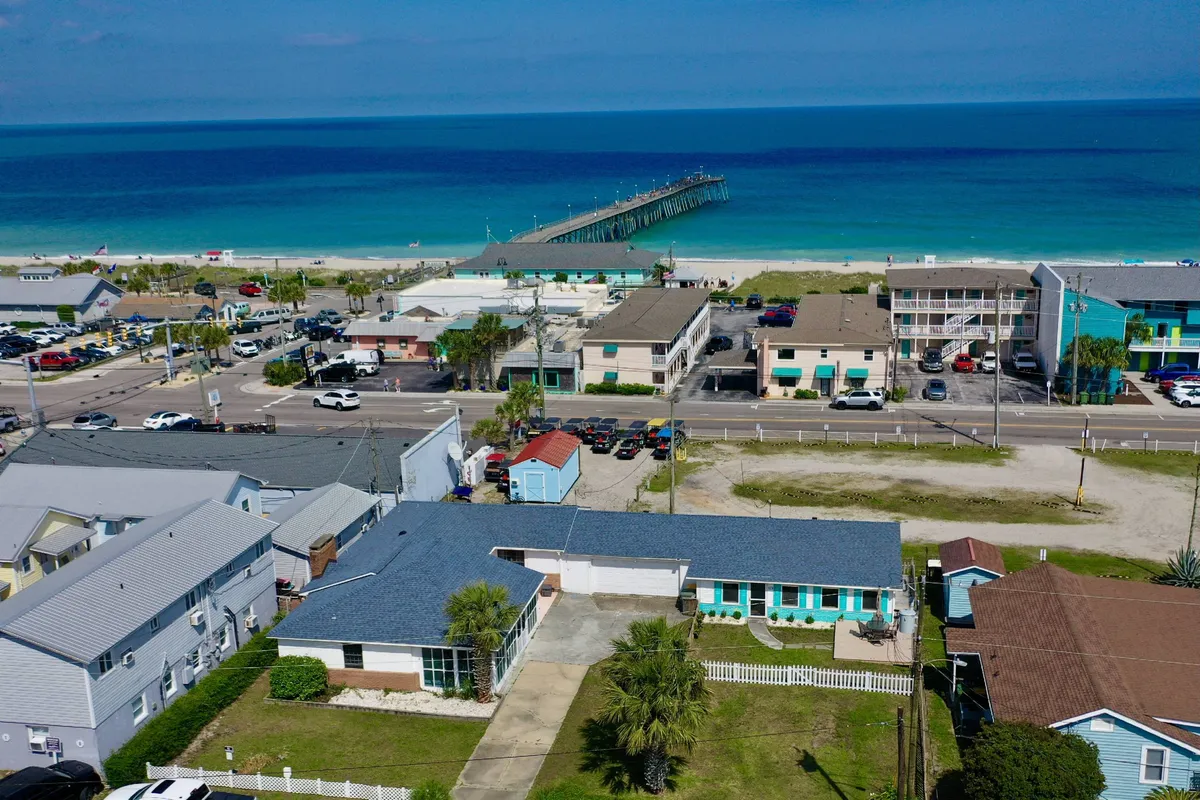 A vibrant exterior view of the Kure Beach shoreline featuring the beachfront, iconic pier, colorful buildings, and palm trees. The scene is lively with visitors engaging in swimming, fishing, and strolling along the sand and pier, conveying a quintessential beach vacation atmosphere.