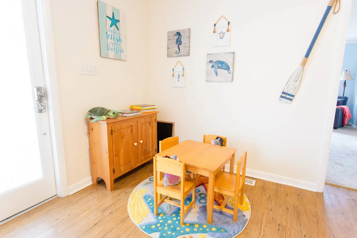 A cozy corner with a wooden table and chairs, colorful rug, decorative wall art, and a wooden cabinet, designed specifically for children’s play. Toys and books are within reach, and the cheerful decor fosters imaginative play and family bonding. The light and colors create a nurturing, inviting mood.