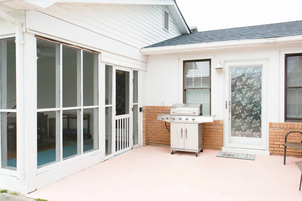 This exterior shot focuses on the gas grill beside the screened porch, set against a brick wall and glass doors. The scene is ready for a barbecue, with space for entertaining guests or family members to gather outdoors.