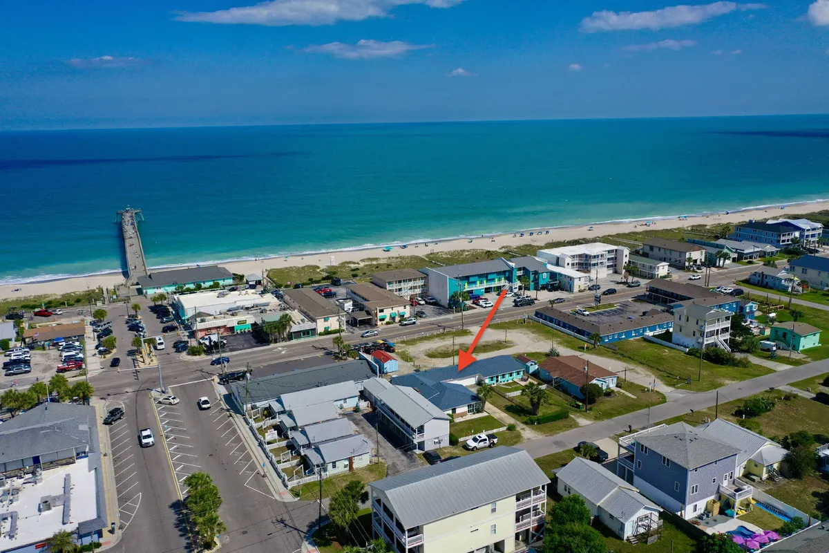 A scenic exterior view featuring a sandy beach, a pier stretching into the ocean, and colorful buildings lining the waterfront. The image also shows a road that gives a sense of the neighborhood's easy access to attractions and the shoreline. Families and visitors can be seen enjoying swimming, fishing, walking, and sunbathing, with the relaxed coastal atmosphere evident throughout.