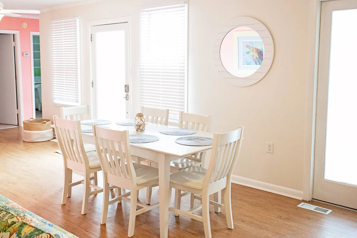 Featuring a spacious dining area with a large table, comfortable chairs, a decorative mirror, and ample natural light streaming through a nearby doorway, this image embodies a friendly and relaxed setting for family meals. The space is open and accessible, with decor elements that evoke a coastal vibe and encourage gatherings at the heart of the home.