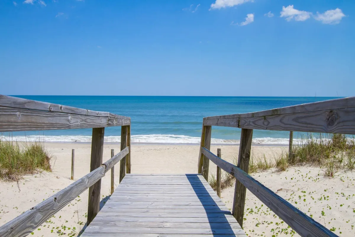 A wooden walkway leads from the property directly toward the ocean, framed by a vast sky. The level pathway underscores the promise of a leisurely, accessible stroll to the beach, inviting relaxation and adventure.