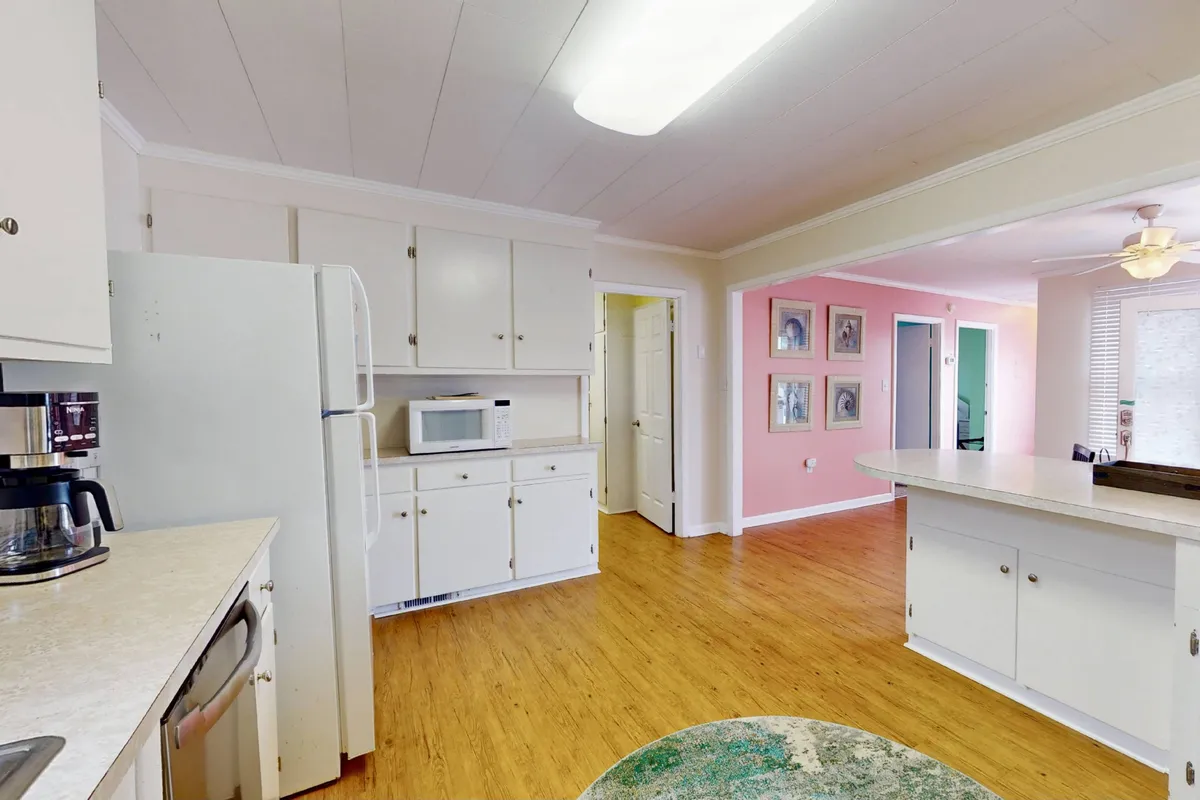A kitchen interior with pristine white cabinetry, a prominent kitchen island, and soft pink walls, evoking a welcoming and comfortable space. The area is arranged for both cooking and casual dining, suggesting versatility for family breakfasts and gatherings.