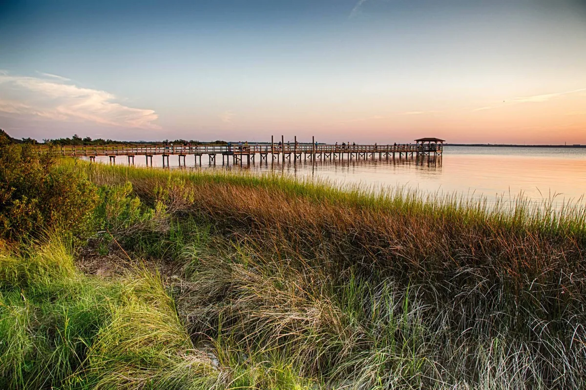 A serene view of a pier stretching over calm water with a grassy shoreline, ideal for fishing, boating, or enjoying sunset moments. The open space accommodates larger groups and invites gatherings or quiet reflection by the water.