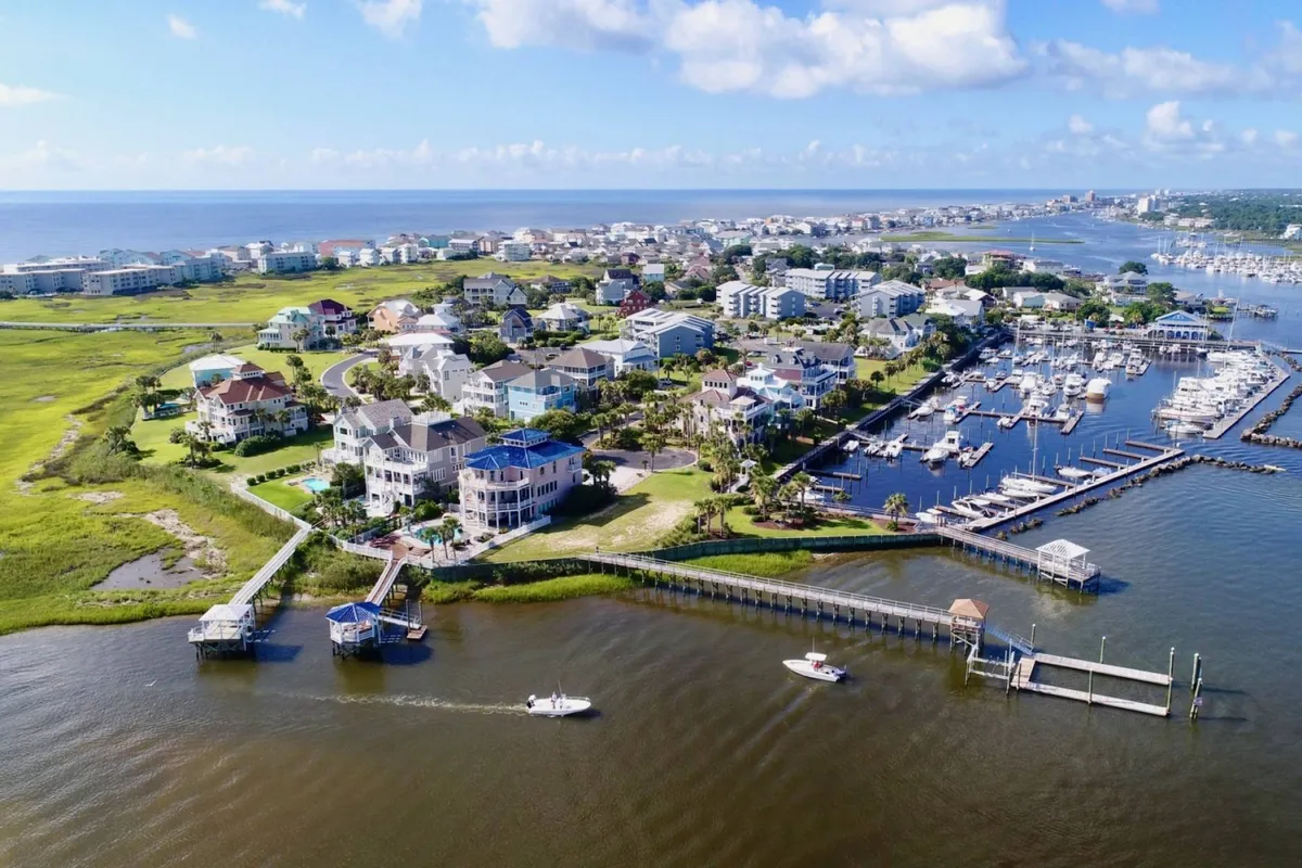 This image shows a peaceful marina scene with residential homes lining the waterways and generous green spaces. The view encompasses boating docks, shimmering water, and areas where families and groups might gather outdoors. The setting suggests local attractions and the inviting coastal neighborhood environment.