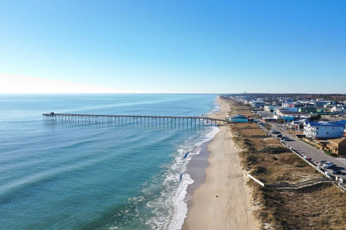 A sweeping view of the iconic pier stretching out over the ocean, with the sandy beach, gentle waves, and the nearby town visible in the background. The scene is vibrant with beachgoers and the lively atmosphere of a coastal hotspot.